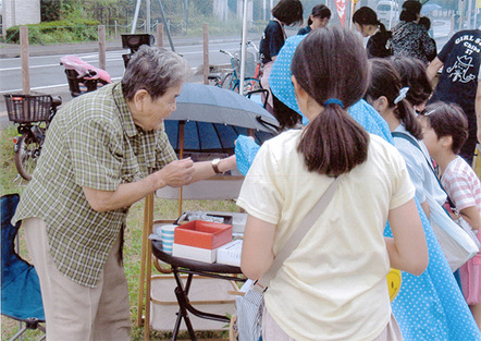 花の精霊夏祭り　四季の会の宣伝活動『花の精霊夏祭り』も4年目、昨年に引き続き団地ふるさと祭り会場に出店しました。オリジナル・グッズを提供して寄付金をお願いするふるさと納税方式、けやき広場ではお目にかかれない方々との交流が魅力です。今年は、鉢物や花のポプリに加えて自治会産花梅の梅ジャムも用意したところ、2時間もしないうちに完売（？）となりました。出店費用を大幅に上回る寄付金2,773円が寄せられています。写真はご主人の遺志を引き継いで活動する世話人Aさんがラベンダーのポプリや特製下敷きを売り込む場面、その抜群の商才に脱帽です（四季の会　7月13日撮影）。